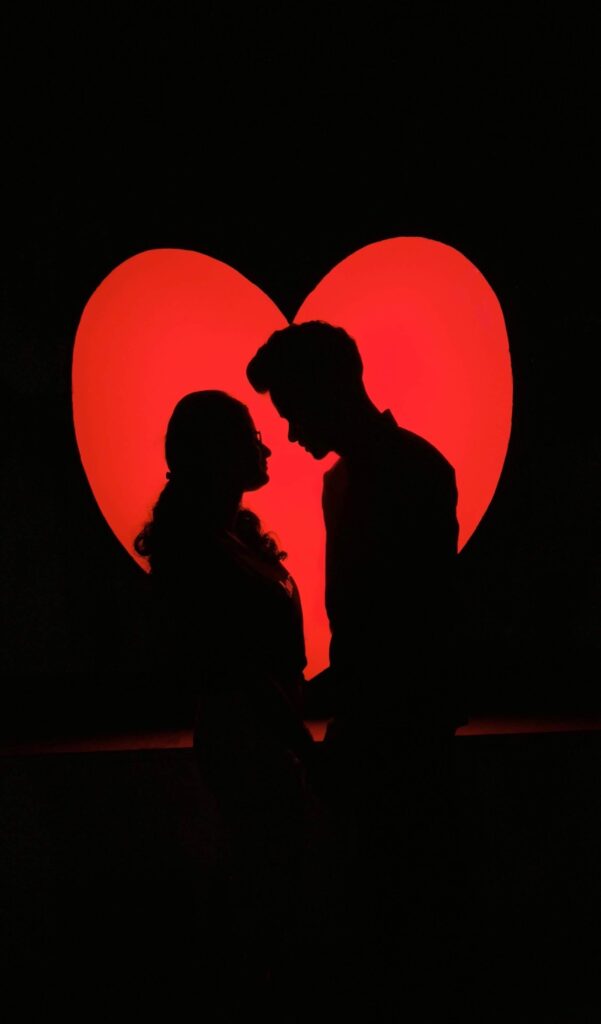 Silhouette of a couple standing close together in front of a glowing red heart, symbolizing romantic love.